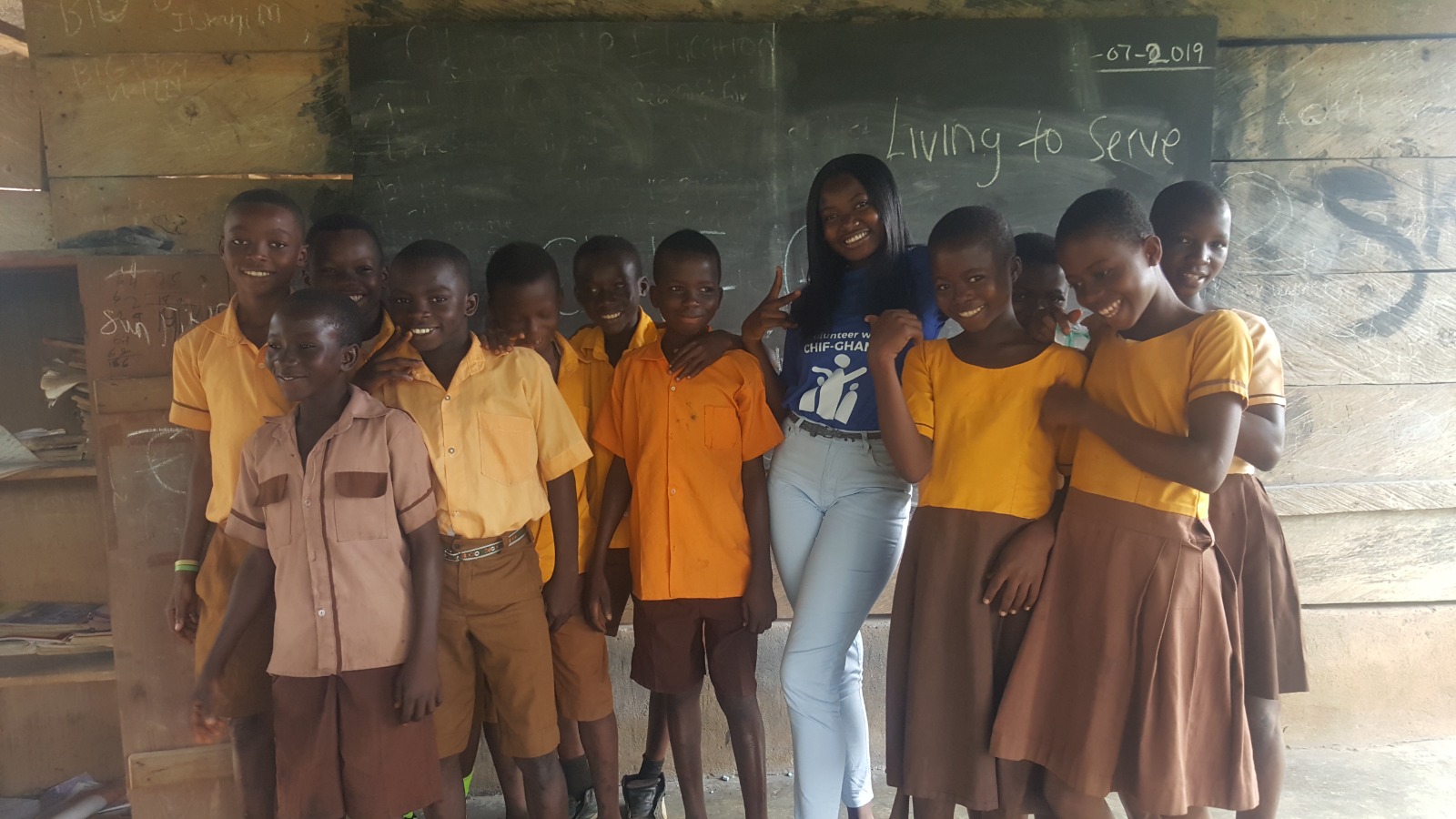 Volunteer smiling with students in front of a chalkboard that reads Living to Serve